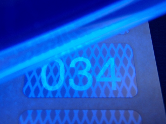 Blaues UV-Licht macht die Sicherheitsnummerierung lesbar Die Sicherheits-Nummerierung auf silberner Rautenmusterfolie ist erst unter blauem UV-Licht sichtbar